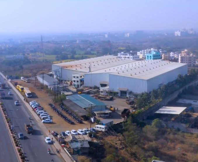 Aerial view of a large white-roofed factory beside a highway, with parked cars and trucks in its yard
